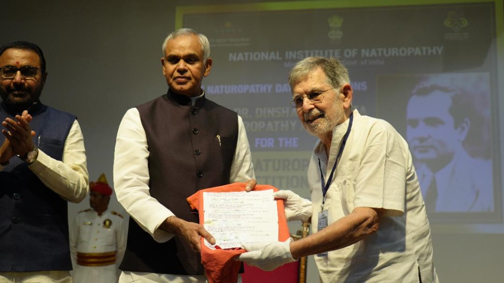 Bahram R. Shahmardaan receiving recognition at the National Institute of Naturopathy, Ministry of AYUSH, Govt. of India, with Dr. Dinshah K. Mehta honoured on screen.